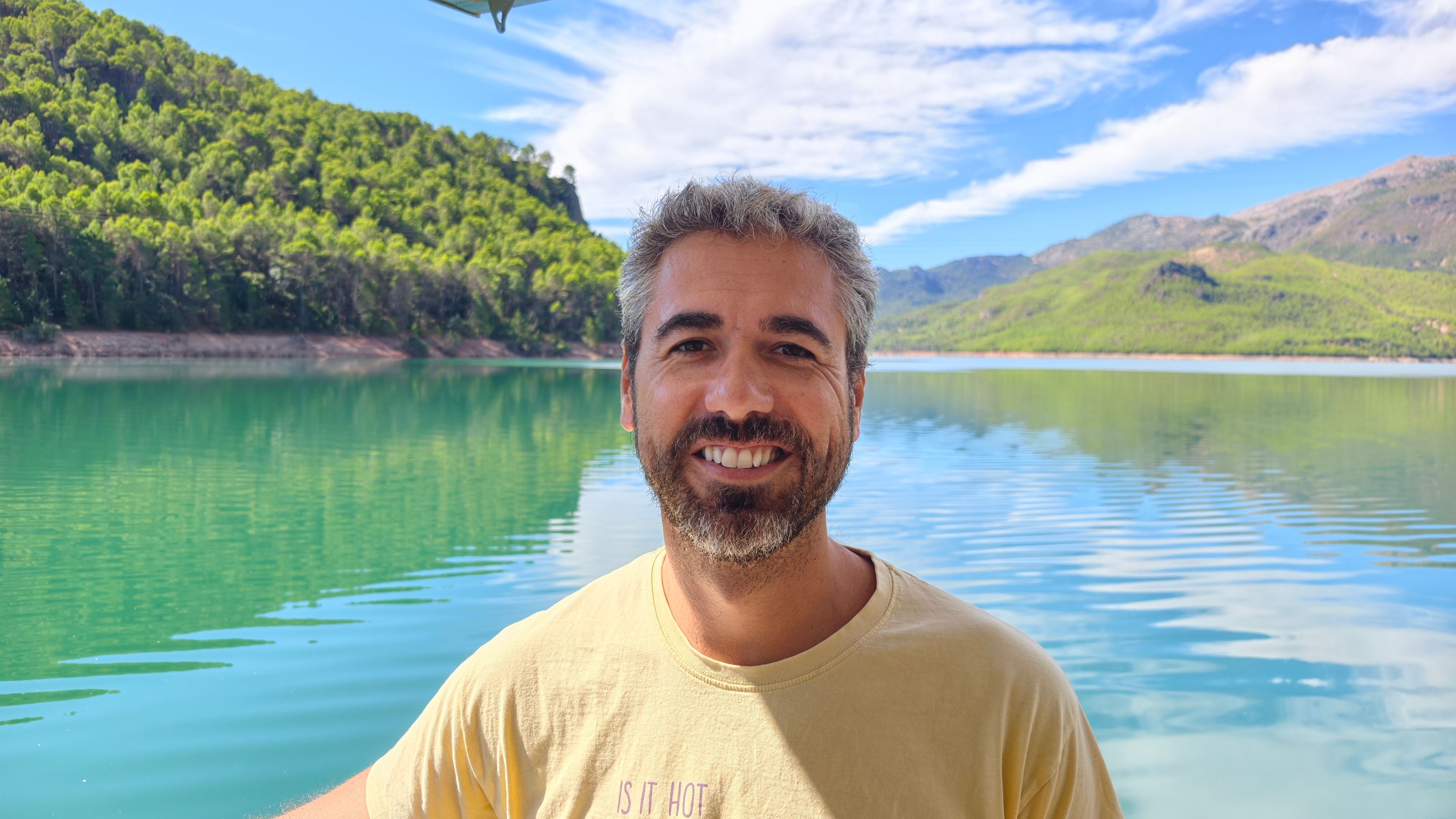 A photo of me posing: a middle-aged man with graying hair and a short beard smiles warmly in the foreground, looking directly at the camera. He is wearing a pale yellow T-shirt. He is on a solar-powered boat on the Tranco Reservoir in Jaén. Behind him, a vast expanse of deep turquoise water stretches into the distance, with gentle ripples on the surface. On both sides of the water, densely forested hills covered in green pine trees rise up, and in the background, taller, rocky mountains are visible. The sky is bright blue with scattered thin white clouds. The lighting is bright, natural sunlight. The photo is taken from a medium-shot perspective, at eye level.